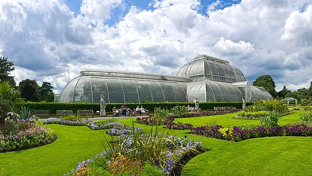 La Palm House di vetro e ferro bianco curvo dietro le aiuole vittoriane e il prato, sotto un cielo di nuvole sparse